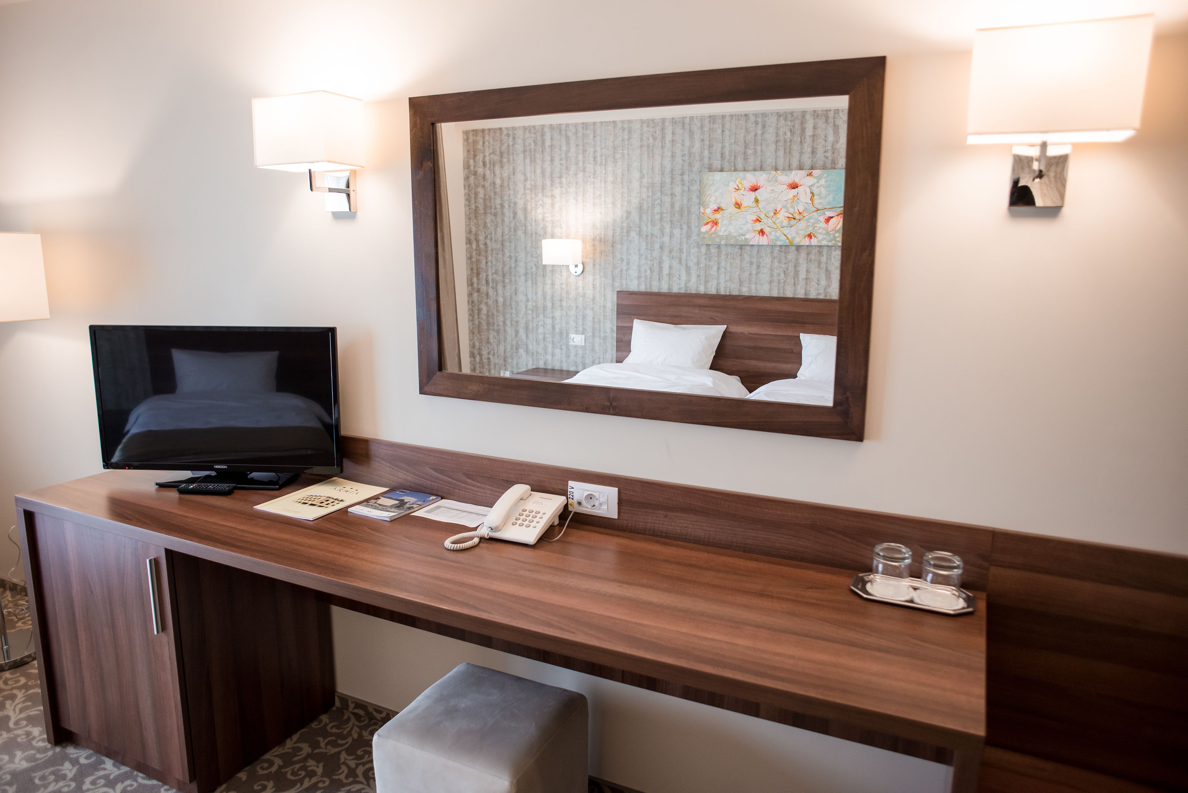 Ett hotellrum med ett skrivbord i trä, en tv, en telefon och en spegel som reflekterar sängen.