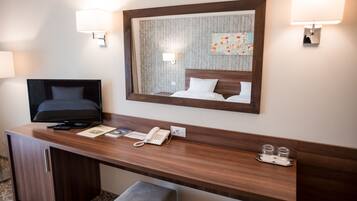 Ett hotellrum med ett skrivbord i trä, en tv, en telefon och en spegel som reflekterar sängen.