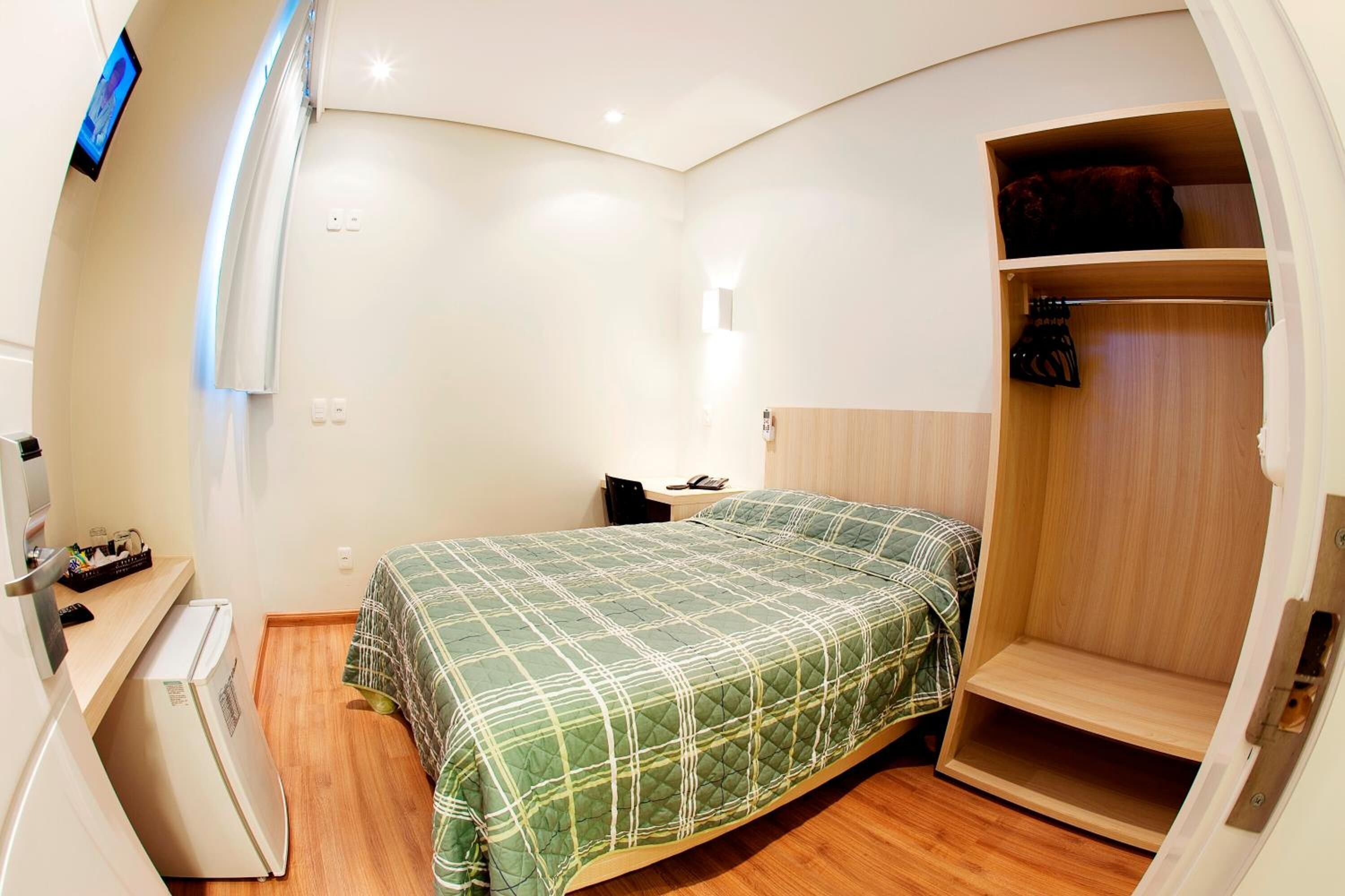 A compact bedroom with a bed, a small desk with a refrigerator, a bedside table, and a wardrobe.