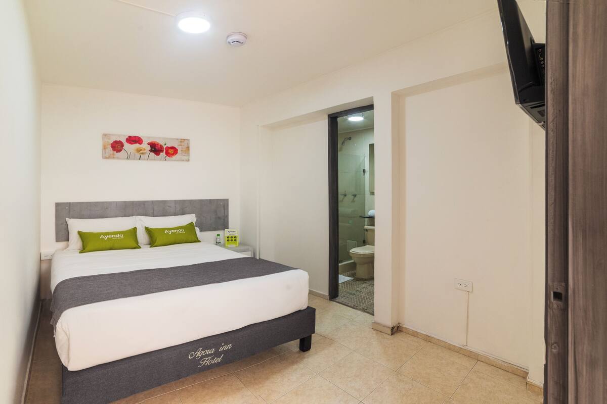 Habitación de hotel con cama, mesita de noche, un cuadro y un baño visible a través de una puerta abierta.