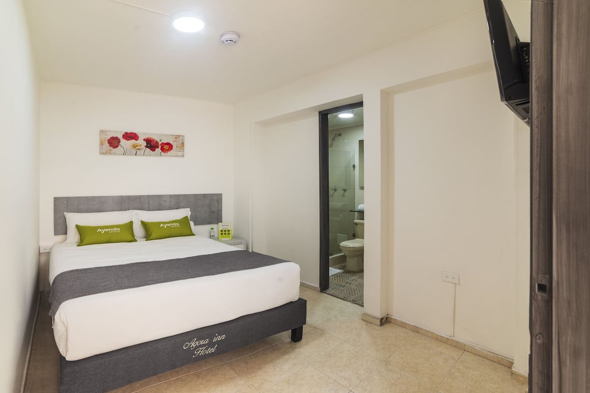 Habitación de hotel con cama, mesita de noche, un cuadro y un baño visible a través de una puerta abierta.