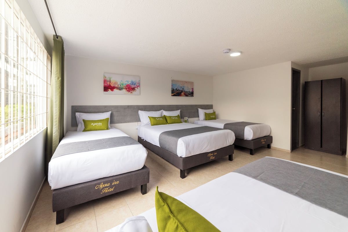 Habitación de hotel con tres camas, cada una con cabecero gris y ropa de cama blanca. Hay dos cuadros en la pared, una ventana con cortina y un armario.