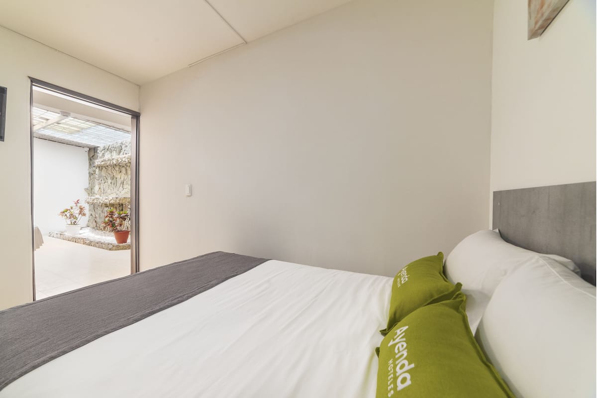 Habitación de hotel con cama, cabecera gris y vista a un balcón con plantas en macetas.
