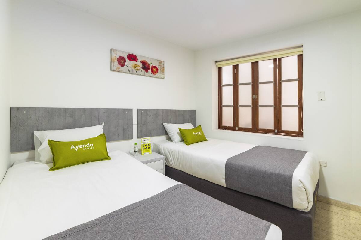 Habitación de hotel con dos camas, cada una con cabecero gris, ropa de cama blanca y almohada verde con el texto 'Ayenda Hotels'. Se ve una ventana con marcos de madera y un cuadro de flores en la pared.