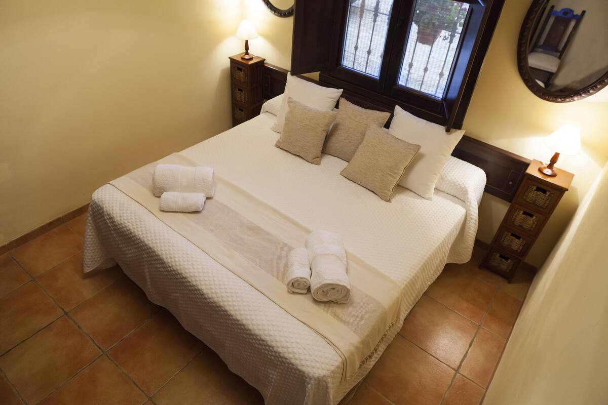 A neatly made bed with white linens and beige pillows, two bedside tables with lamps, a window with a view, and a mirror on the wall.