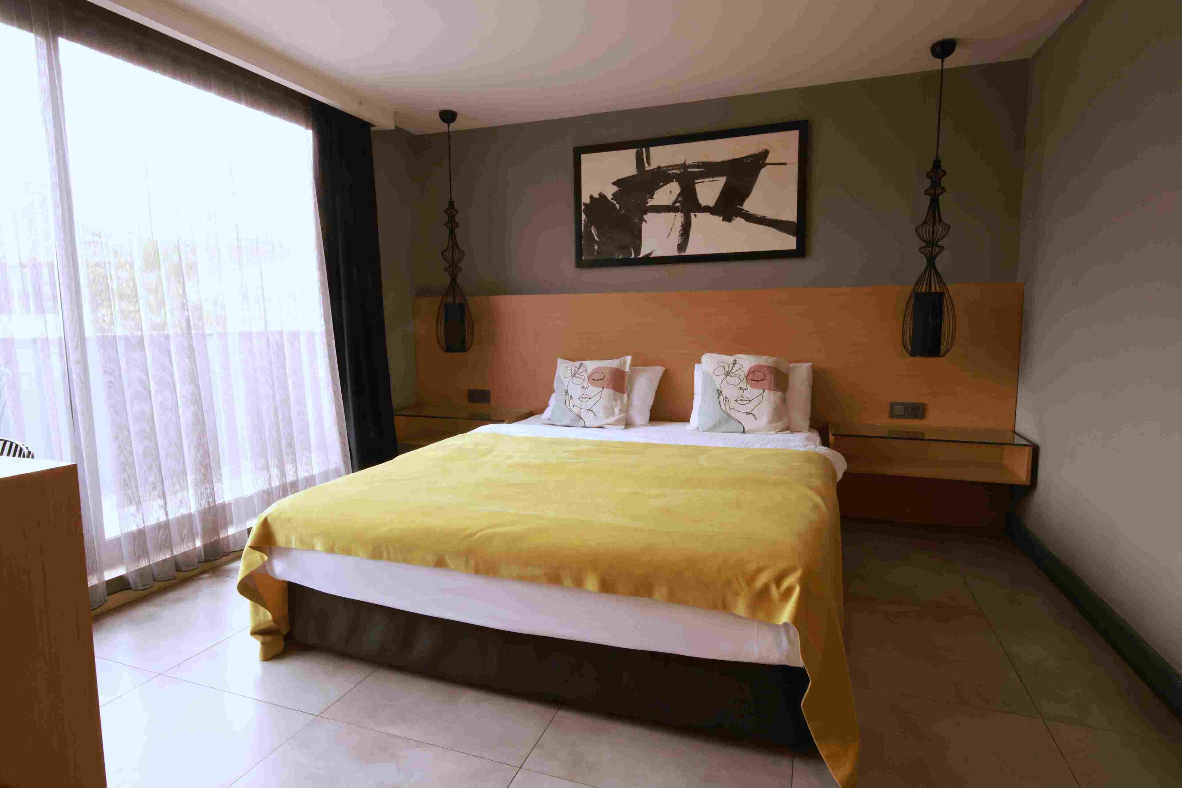 Una habitación de hotel con una cama grande, una mesita de noche, una obra de arte en la pared y una ventana con cortinas.