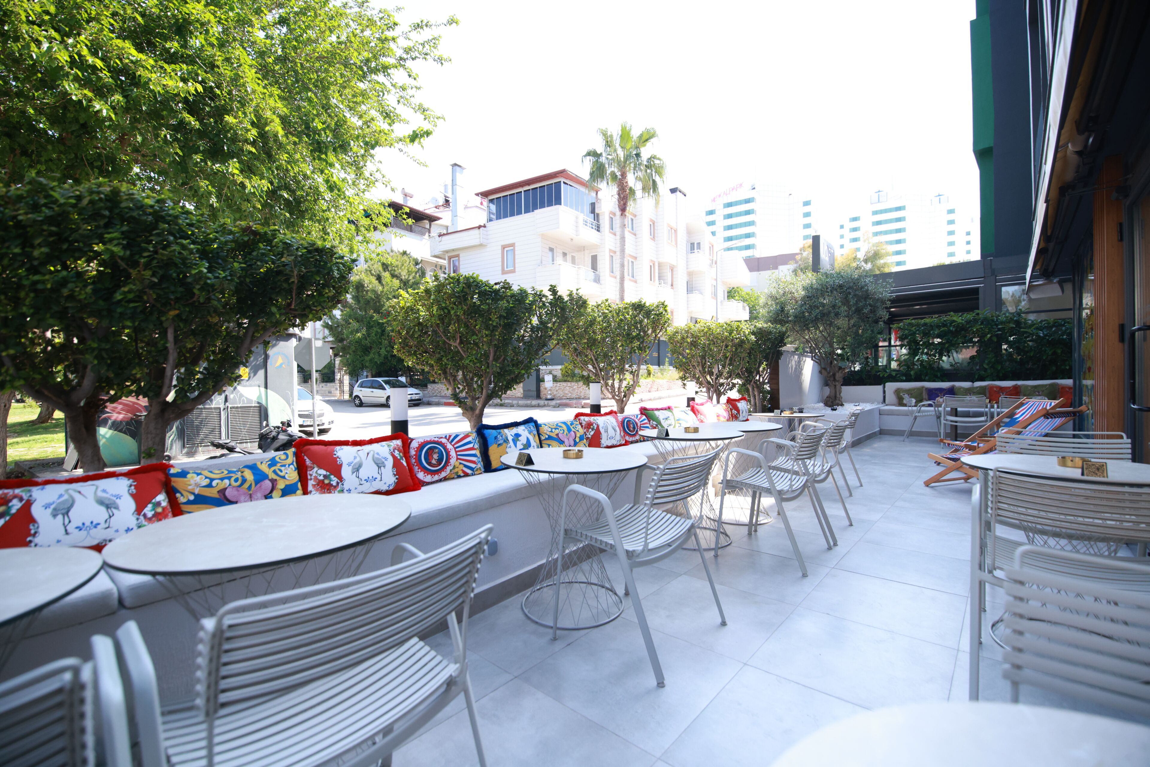 An outdoor cafe area with white metal chairs and tables, a low wall with colorful cushions, and a view of buildings in the background.