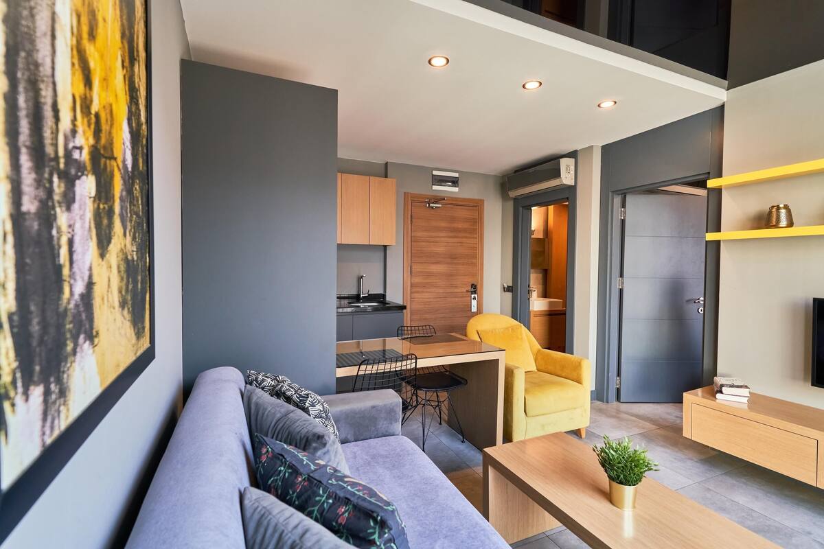Una sala de estar moderna con un sofá gris, un sillón amarillo y una mesa de centro de madera. Se ve una cocina al fondo, con una puerta de madera y una pequeña isla.