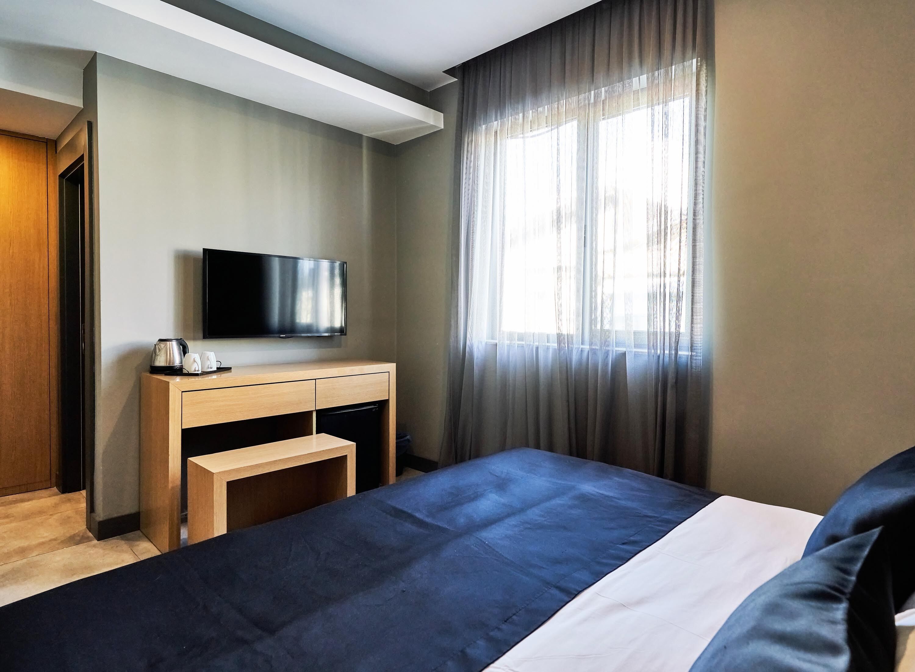 A modern hotel room with a bed, a TV mounted on the wall, a wooden desk with a kettle and cups, and a large window with sheer curtains.
