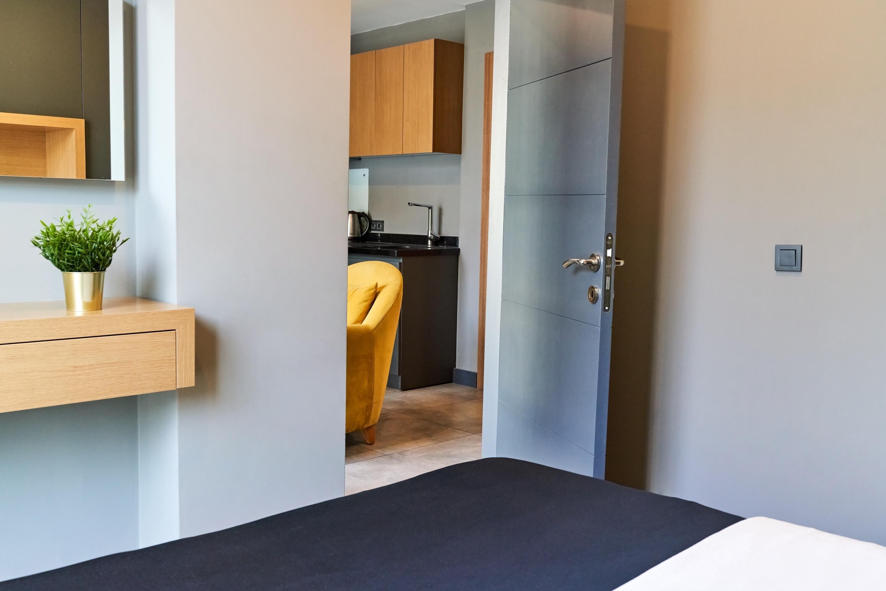 A modern hotel room with a bed, a small bedside table with a plant, and a view into the kitchen area with wooden cabinets and a yellow chair.