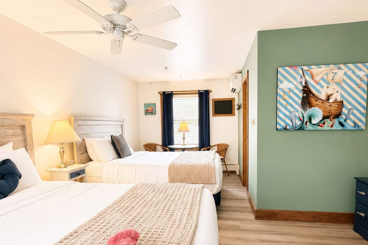 Habitación de hotel con cama, ventilador de techo, ventana con cortinas, un cuadro en la pared y piso de madera.