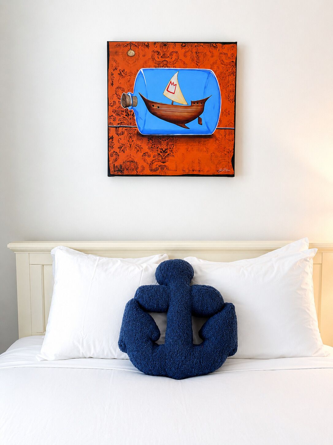 A bed with white linens and a blue pillow shaped like a knot. Above the bed, there is a framed artwork depicting a boat on an orange background.