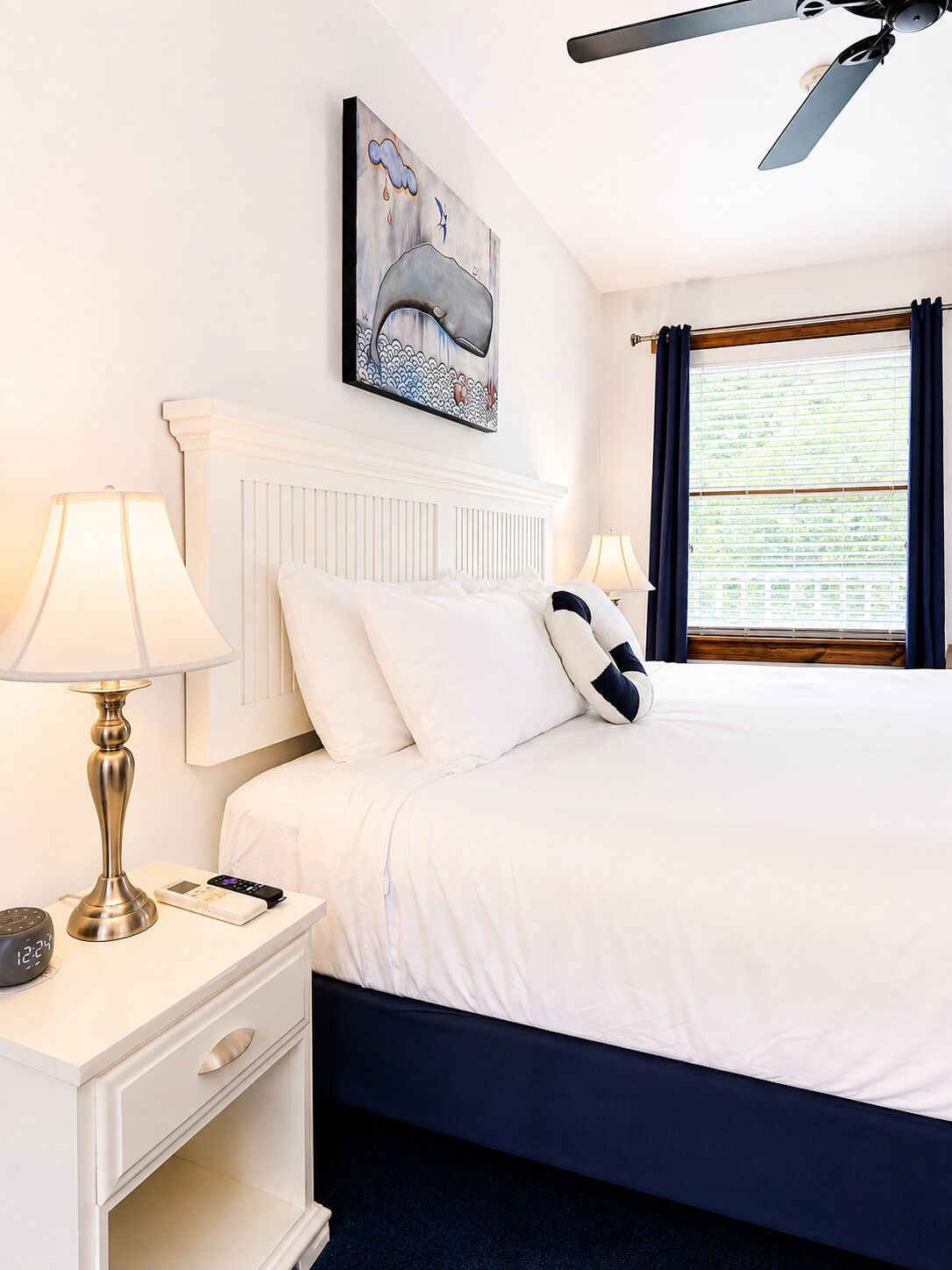 A hotel room with a bed, a nightstand, a lamp, a ceiling fan, a window with curtains, and a painting on the wall.