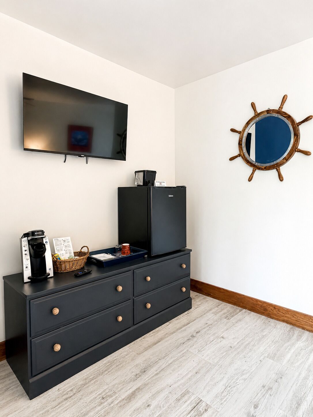 Una habitación moderna con televisión de pantalla plana, cafetera, cesta y un adorno con timón de barco en la pared.