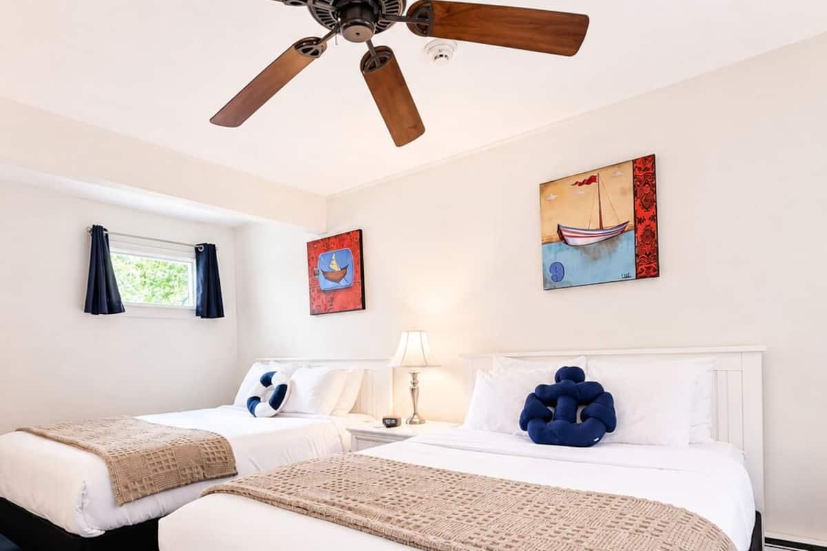 Habitación de hotel con dos camas, ventilador de techo, obras de arte en las paredes, ventana con cortinas y una mesita de noche con lámpara.