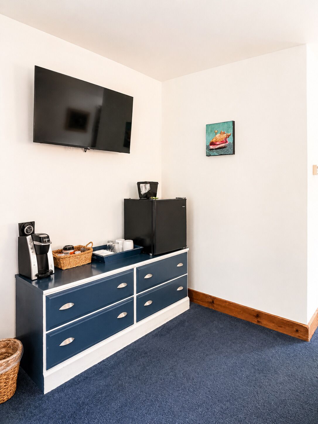 Ein Zimmer mit einem an der Wand befestigten Flachbildfernseher, einem schwarzen Kühlschrank, einer blauen und weißen Kommode mit Schubladen, einem Korbwägelchen und einem kleinen Bild an der Wand.