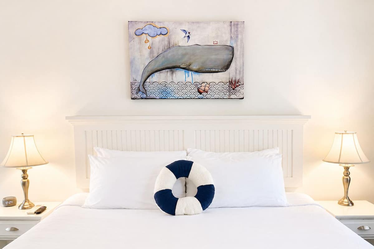Habitación de hotel con cama, dos mesitas de noche, lámparas, un reloj y una obra de arte con temática de ballenas en la pared.