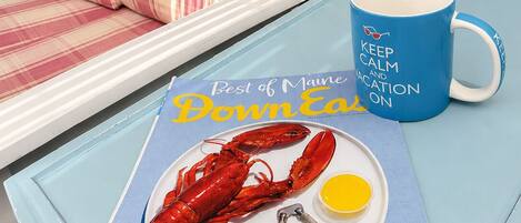 A dining table with a lobster, lemon, and utensils, a blue mug with text, and a plaid sofa in a room with a window and kitchen in the background.