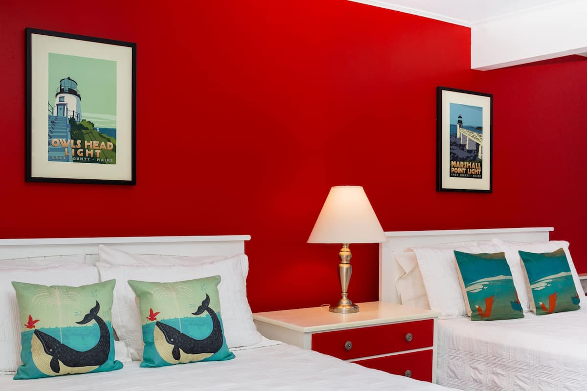 Habitación de hotel con pared roja, dos camas, una mesita de noche con lámpara y dos cuadros con imágenes de faros.