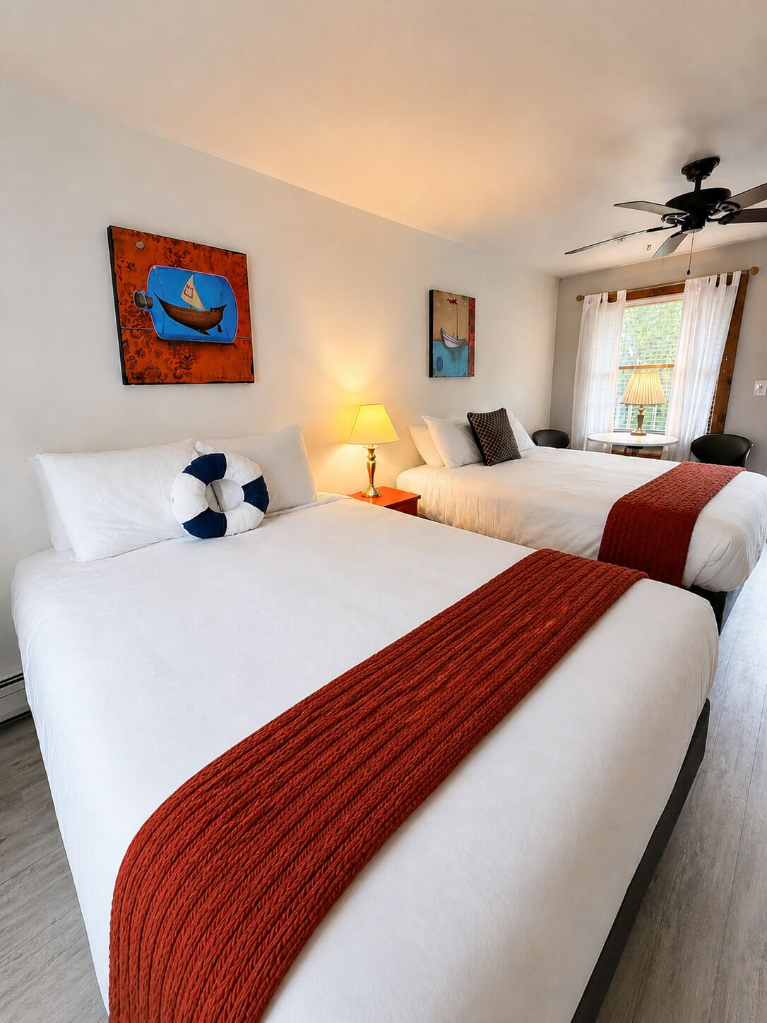 A hotel room with two beds, a ceiling fan, a window with curtains, and artwork on the walls.