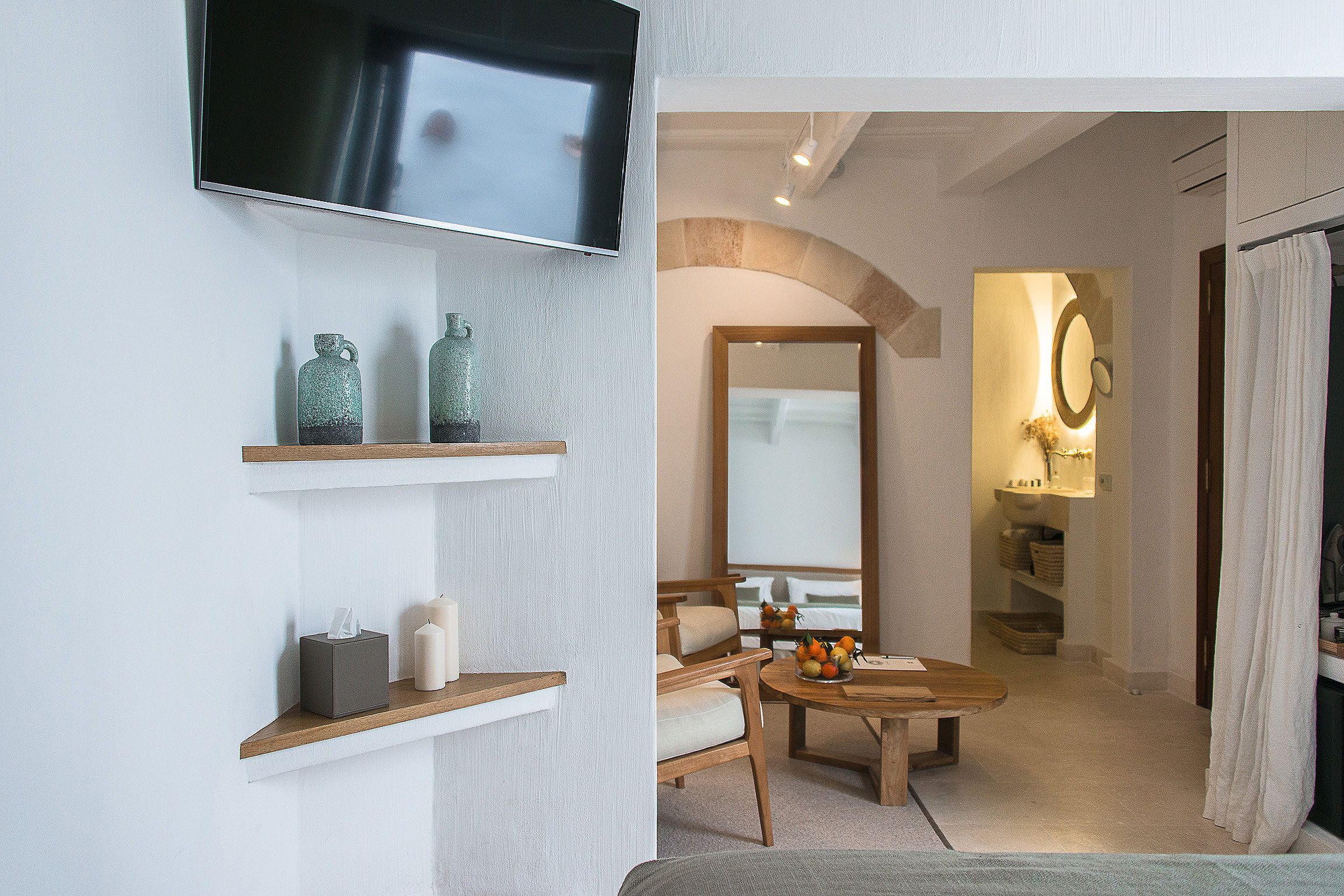 Une chambre d’hôtel moderne dotée d’une télévision à écran plat, d’étagères en bois ornées d’objets décoratifs, d’un coin salon confortable avec une table et d’une salle de bain avec un miroir et un porte-serviettes.