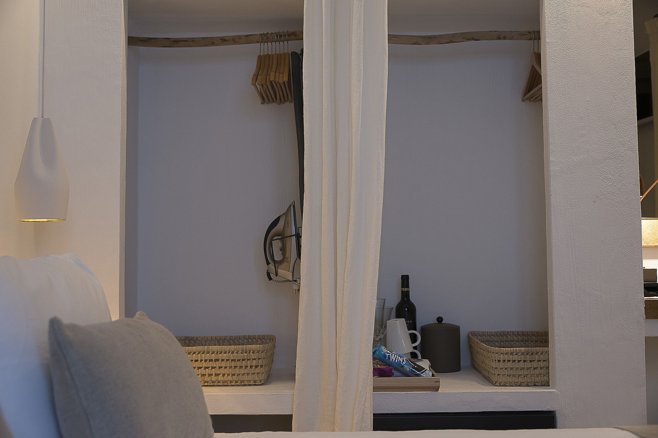 Una stanza con un letto, un comodino, una lampada a sospensione, una finestra vista e uno scaffale con cesti e una bottiglia.