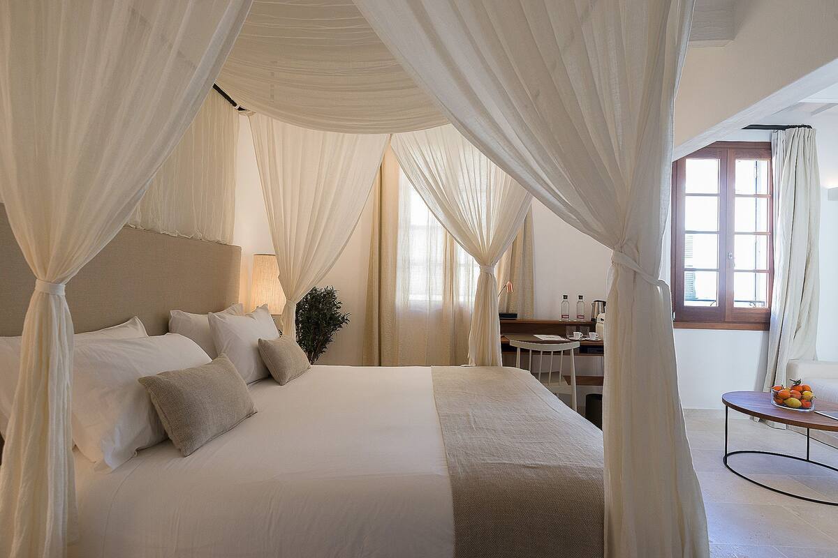 A bedroom with a large bed, white curtains, and a small table with fruit.