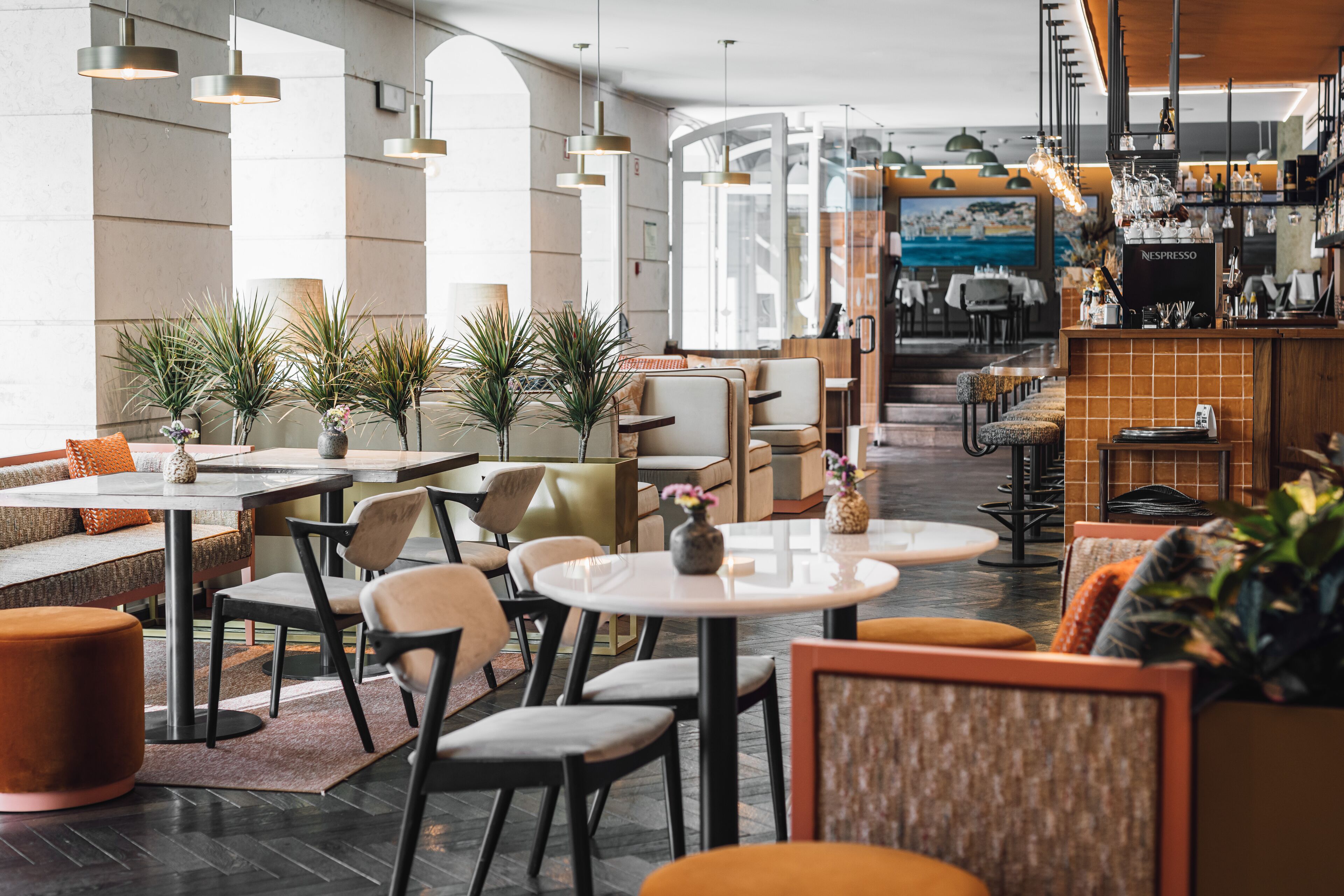 En moderne restaurant med runde borde, polstrede siddepladser, et barområde med en Nespresso-maskine og potteplanter.