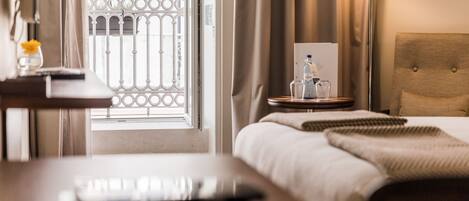 A hotel room with a bed, bedside table, lamp, and a view of the outside through a window with a metal gate.