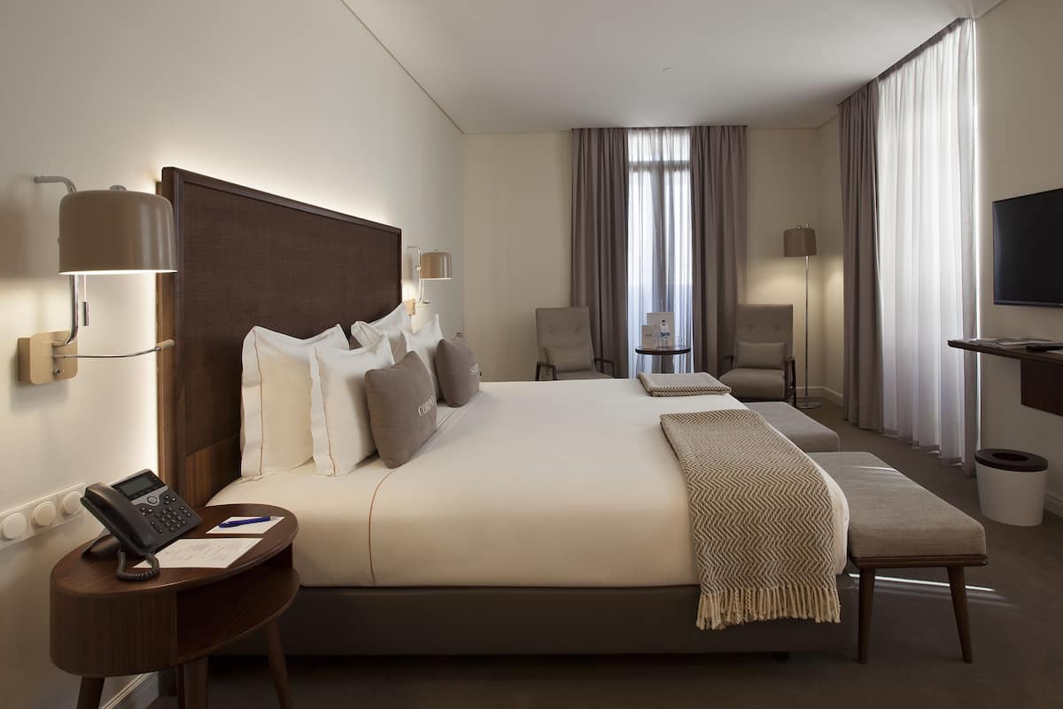 A hotel room with a large bed, bedside tables, a desk, a chair, a TV, and a window with curtains.