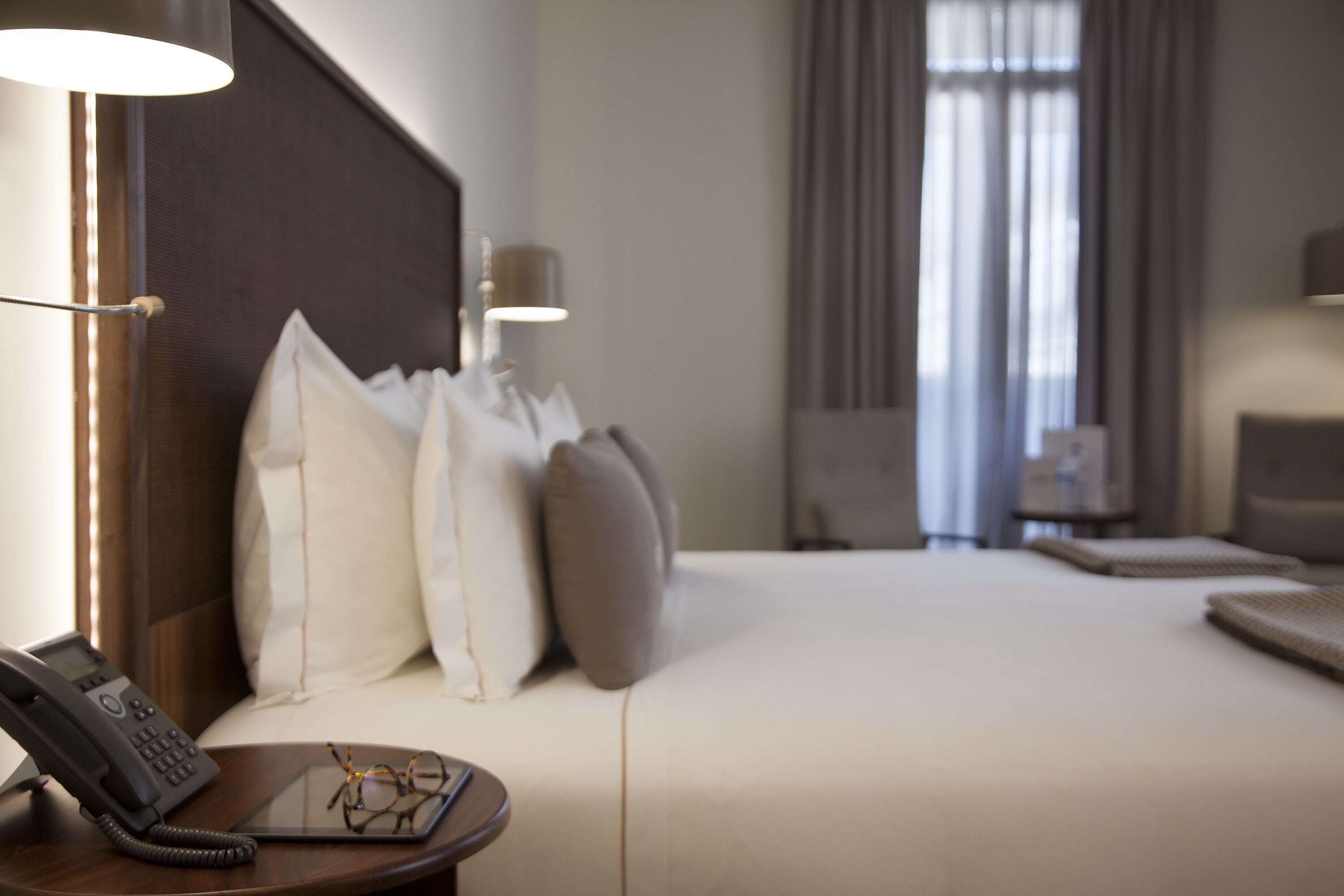 Una habitación de hotel con cama, lámparas de noche, un teléfono y un par de gafas sobre una mesita de noche.