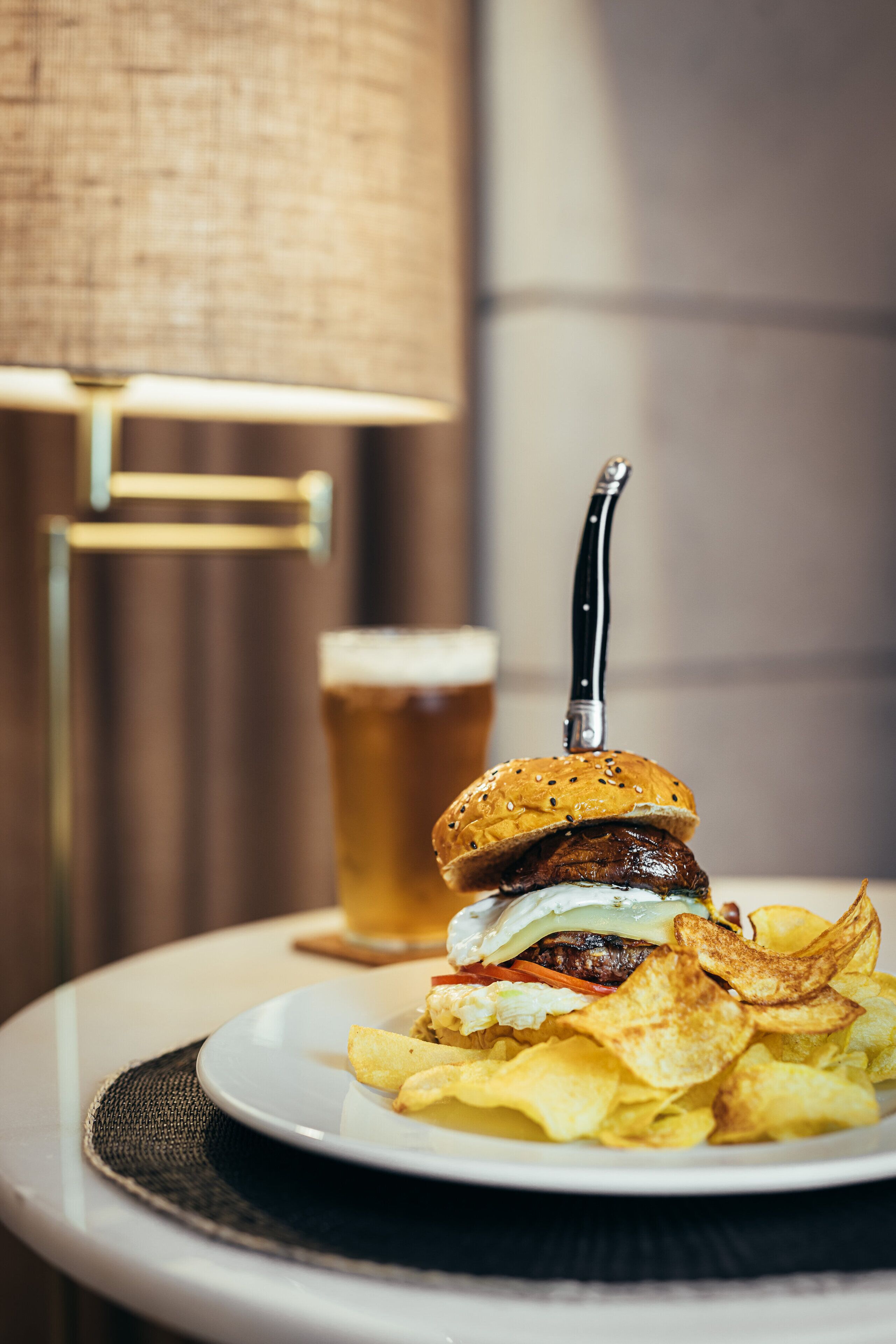 A plate with a burger, chips, and a beer glass on a table with a lamp in the background.