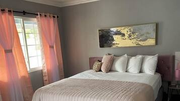Un dormitorio con una cama grande, cabecera, un cuadro en la pared y una ventana con cortinas.