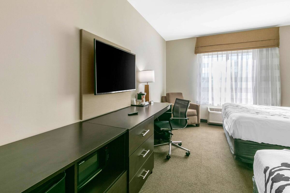 Ett hotellrum med en säng, ett skrivbord med en platt-TV, en stol och ett fönster med tunna, genomskinliga gardiner.