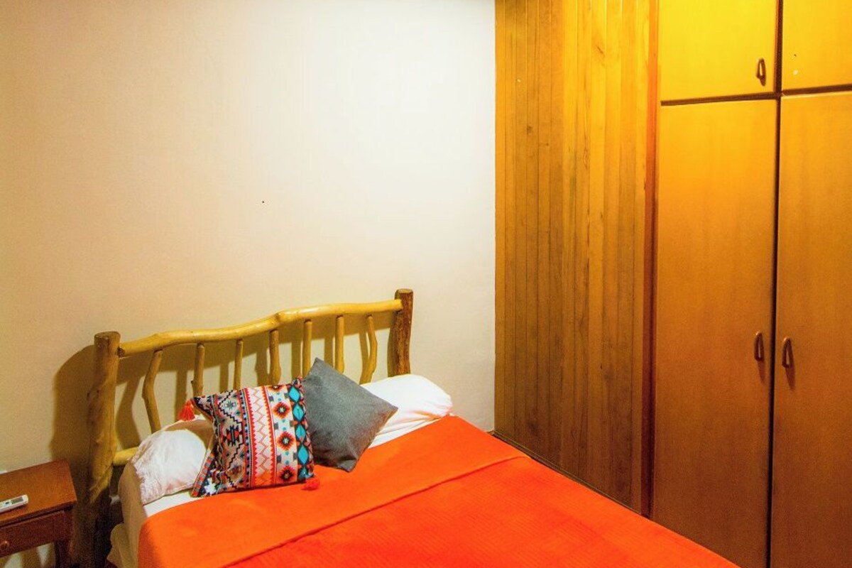 Een slaapkamer met een houten plafond, een bed met oranje sprei, een houten hoofdeinde, een nachtkastje en een garderobe.