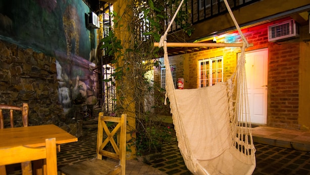 A cozy outdoor seating area with a wooden table and chairs, a hanging hammock, and a stone wall with a mural. - Posada los Tajibos (Puerto Iguazú)