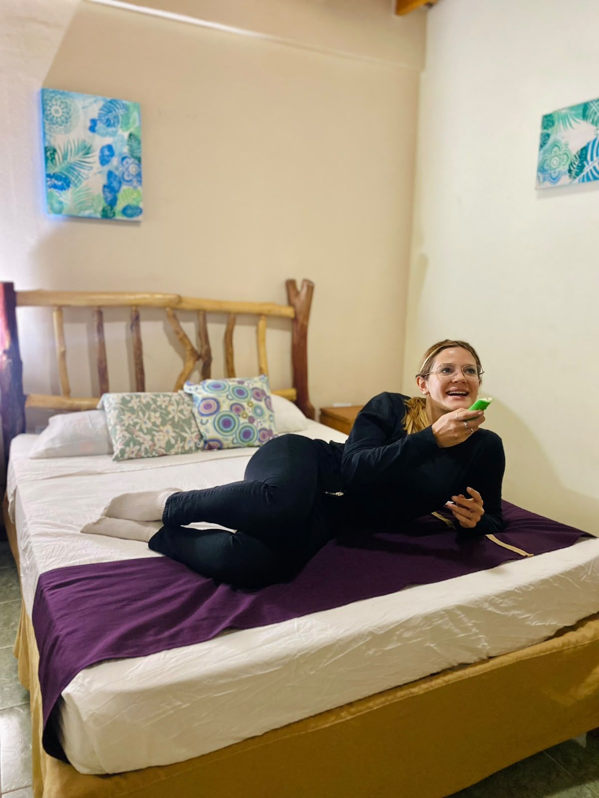 A person lying on a bed with a purple blanket, a wooden headboard, and patterned pillows.