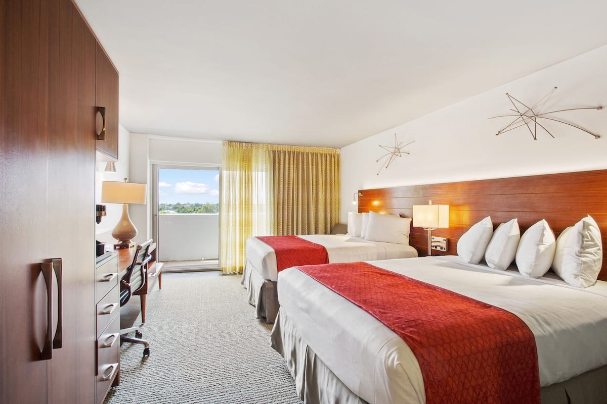 A hotel room with two beds, a desk, a chair, a lamp, and a view of the outdoors.