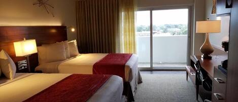A hotel room with two beds, a desk, a chair, a lamp, and a view of the outdoors.