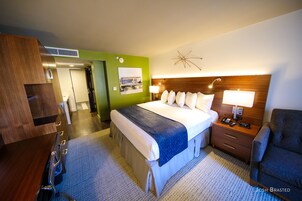 Quarto de hotel com cama, mesa de cabeceira, luminária, cadeira e parede verde.