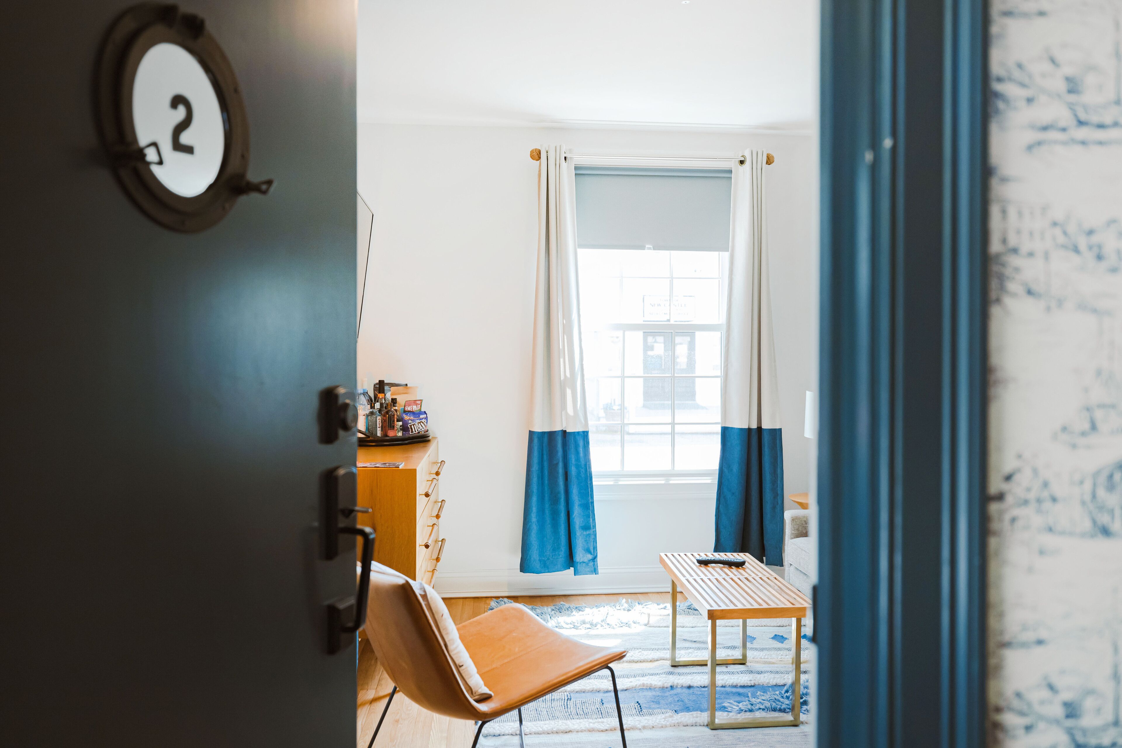 Una habitación con una silla de madera, una mesita pequeña, una ventana con cortinas azules y una placa con número fijada a la pared.