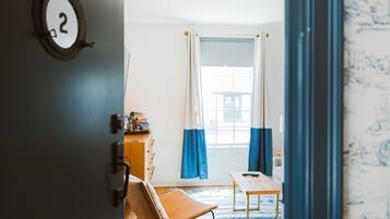 Una habitación con una silla de madera, una mesita pequeña, una ventana con cortinas azules y una placa con número fijada a la pared.