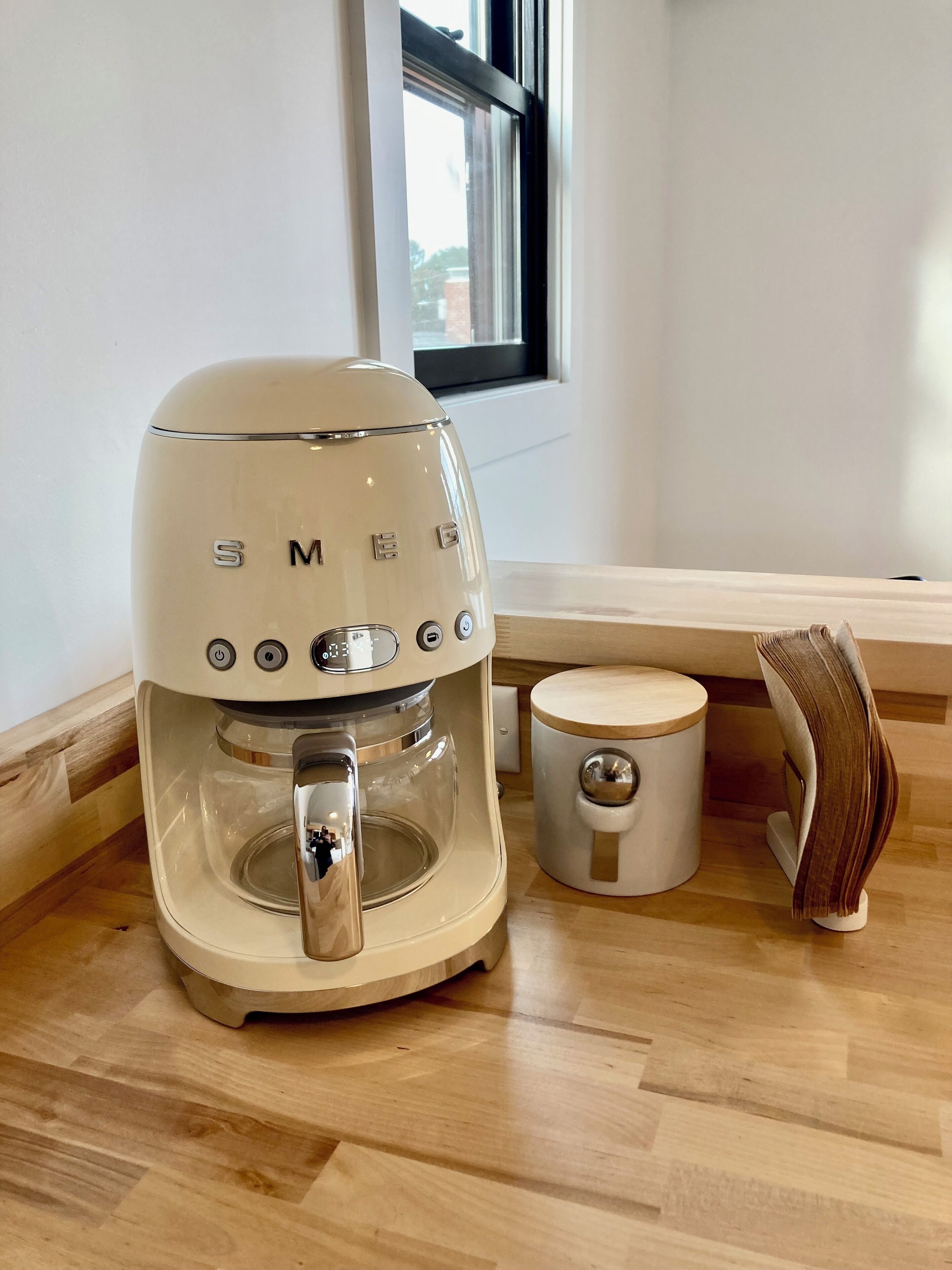 Een koffiezetapparaat met een transparante waterreservoir, een kleine witte mok, en een stapel boeken op een houten tafel.