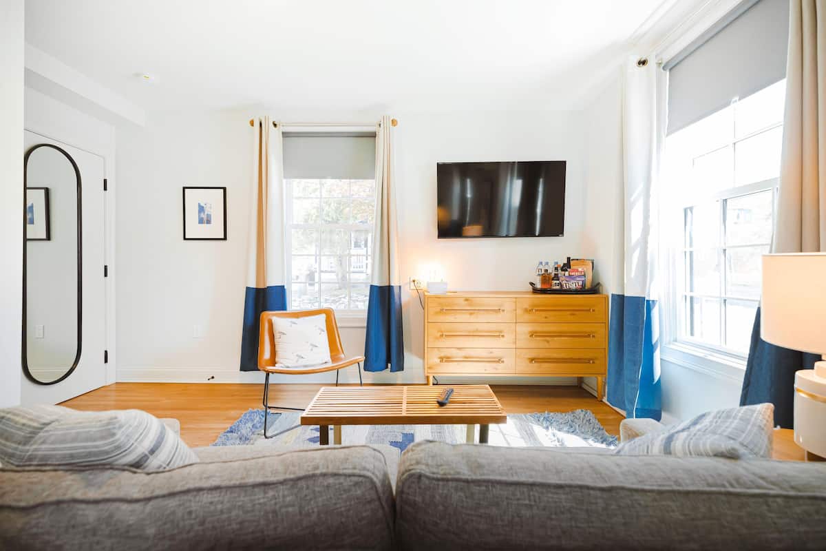 Ein modernes Wohnzimmer mit einer Couch, einem Holztisch, einem Sessel, einem an der Wand montierten Fernseher, einer Holzkommode und einem Fenster mit Vorhängen.