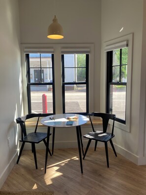 A room with a round table, two chairs, a pendant light, and large windows with a view of the street. - Great Island Inn (New Castle)
