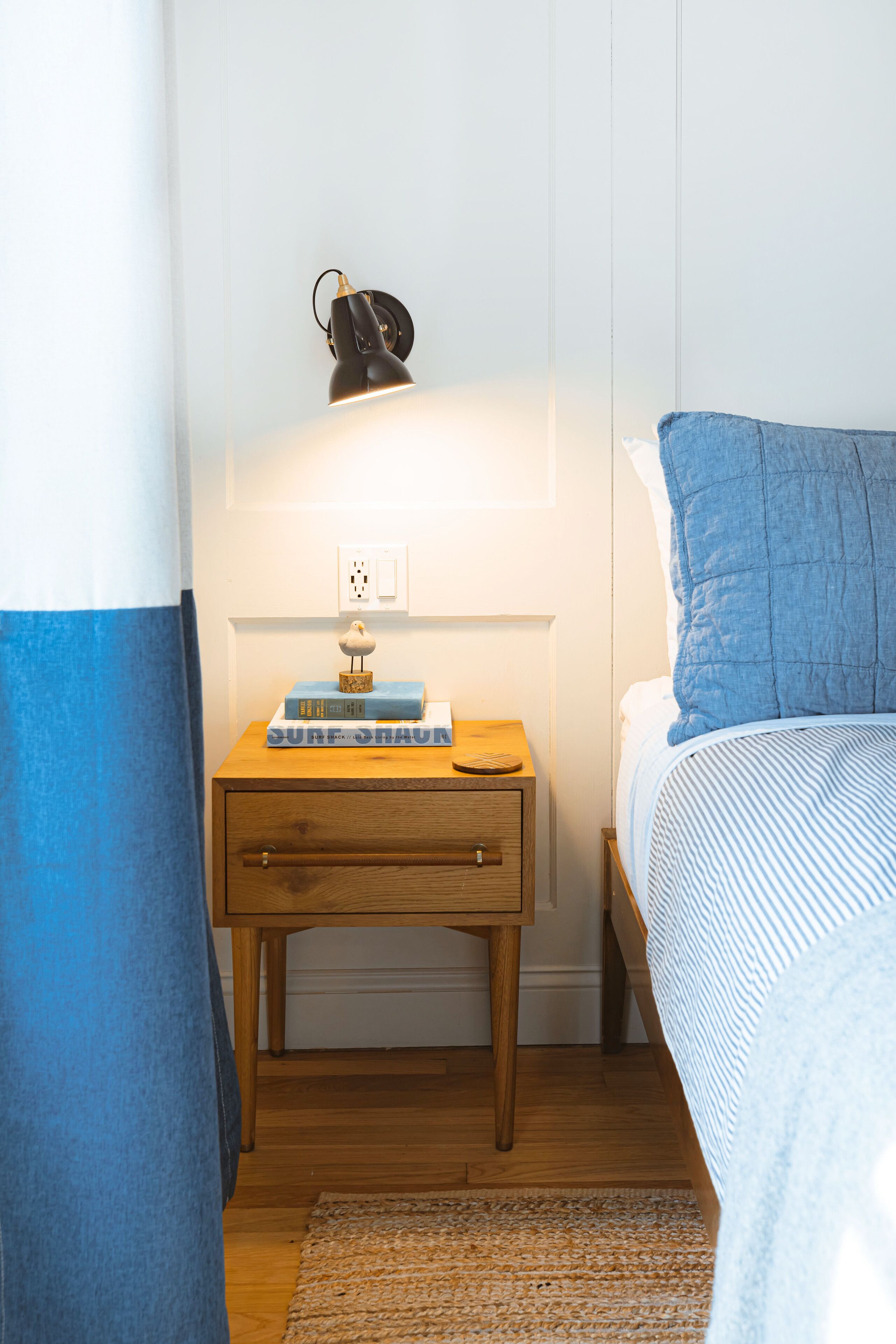 A bedroom with a wooden nightstand, a lamp, a stack of books, and a bed with a blue blanket.