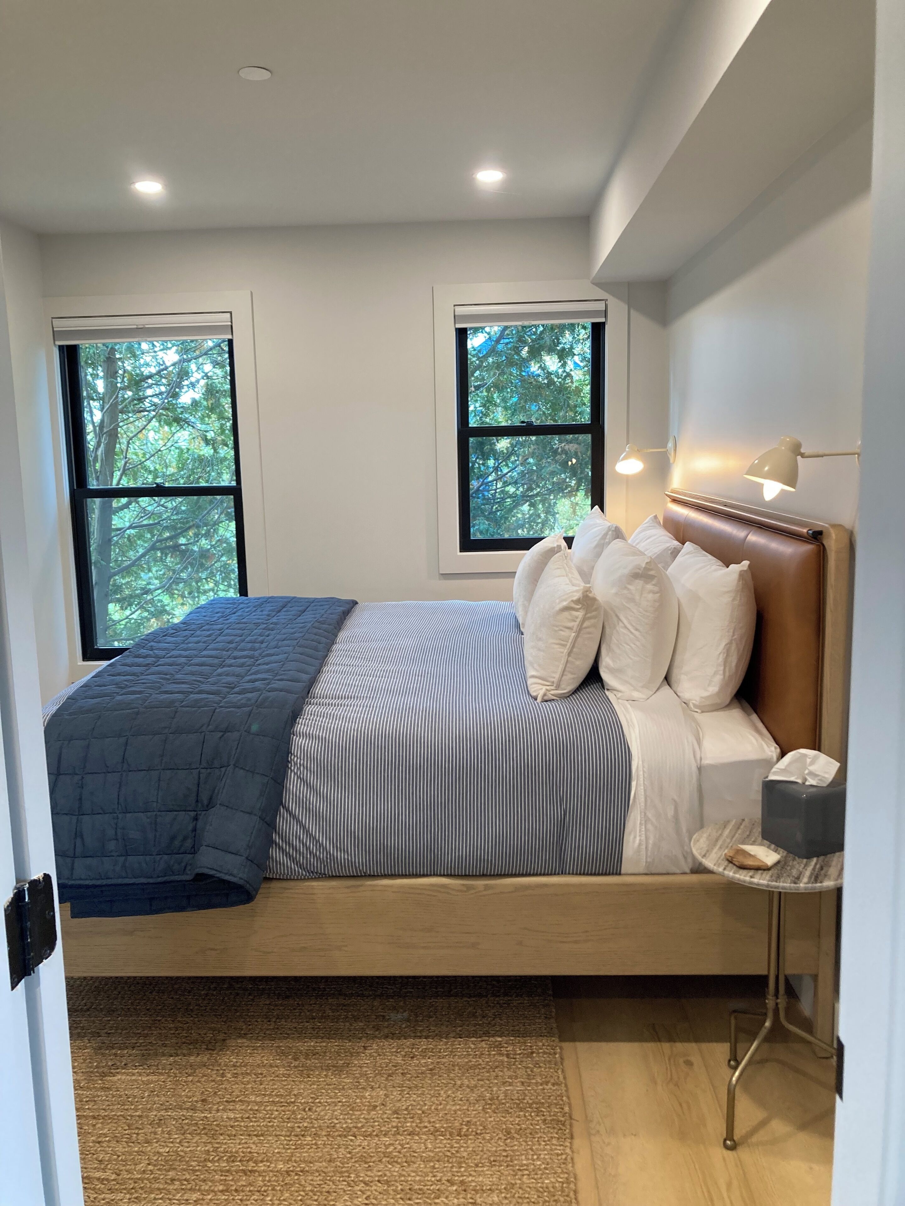 A bedroom with a bed, two windows, a bedside table, and a lamp.