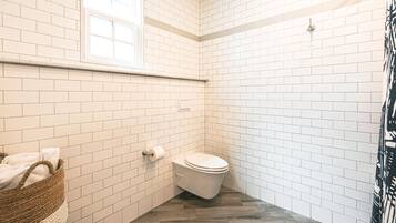 Un baño con paredes de azulejos blancos, suelo de madera, inodoro, ventana y una cesta con toallas.