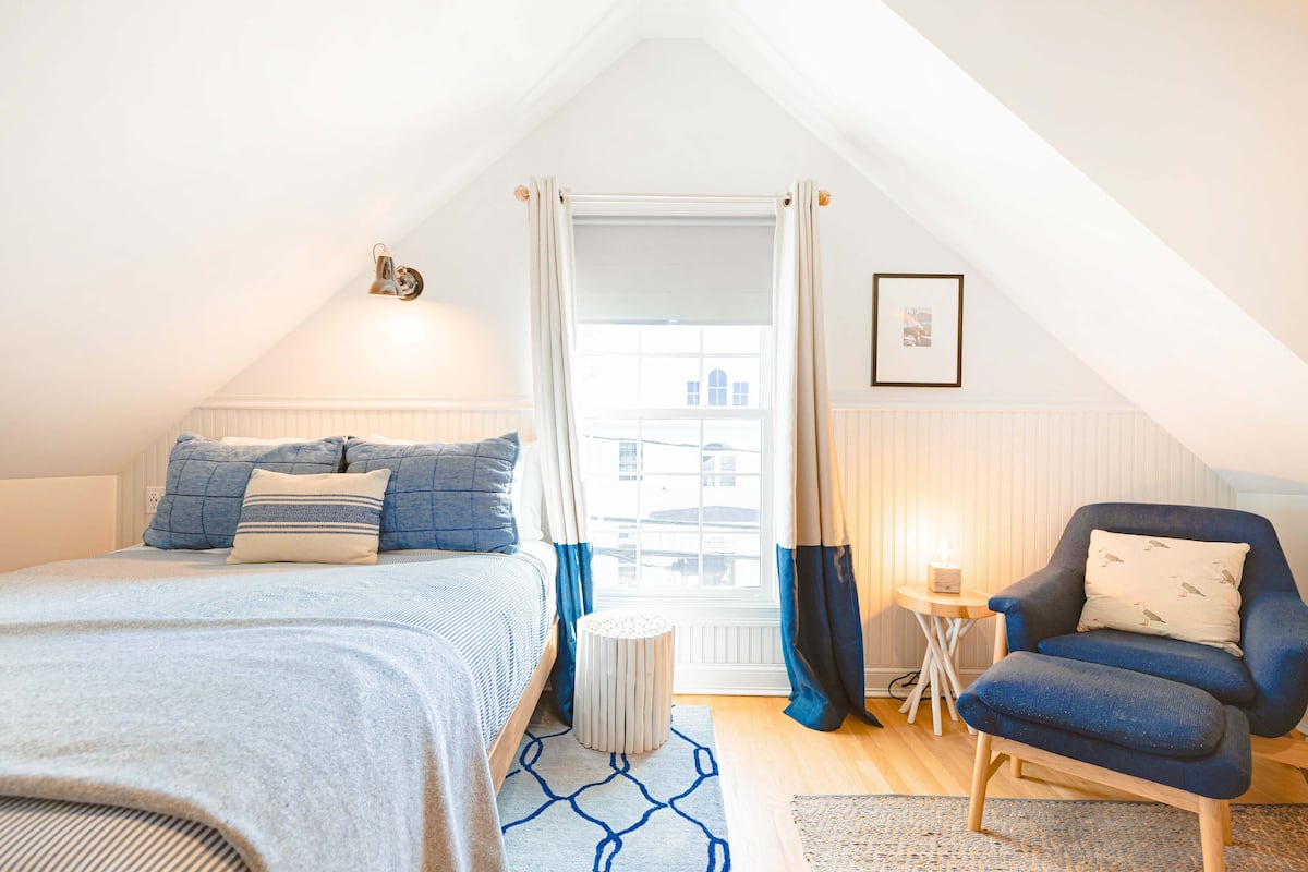 Ein Schlafzimmer mit einem Bett, einem Sessel, einem kleinen Tisch und einem Fenster mit Vorhängen.