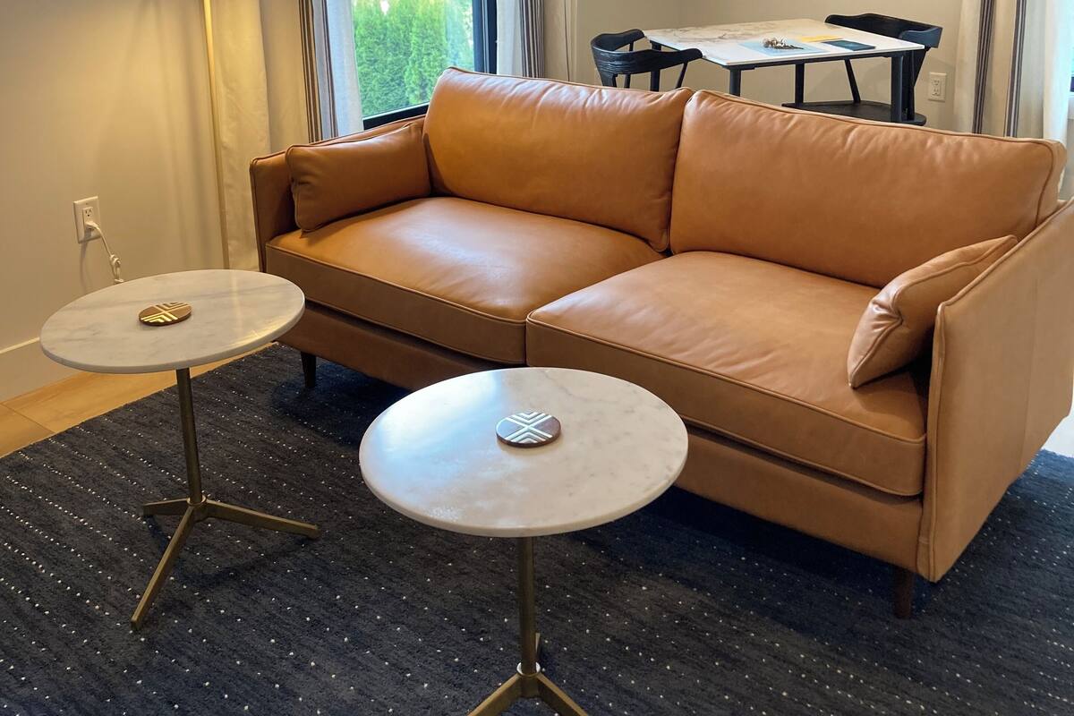 Ein modernes Wohnzimmer mit einem beigen Ledersofa, zwei kleinen runden Tischen mit dekorativen Mittelstücken, einem dunklen Teppich und einer Essecke im Hintergrund.